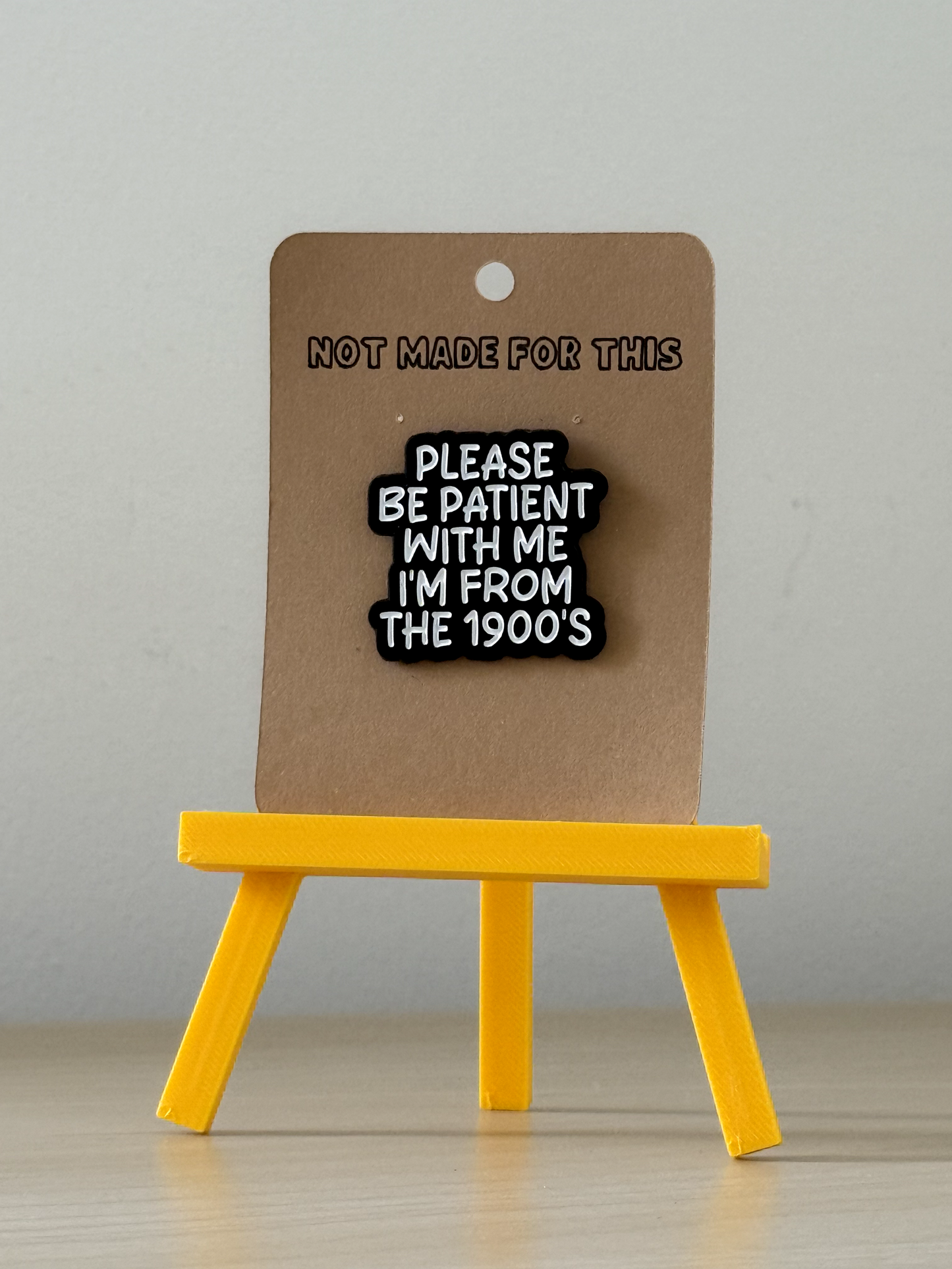 Pin with text 'Please be patient with me I'm from the 1900's' on a brown card with 'Not made for this' text, displayed on a yellow stand against a gray background.