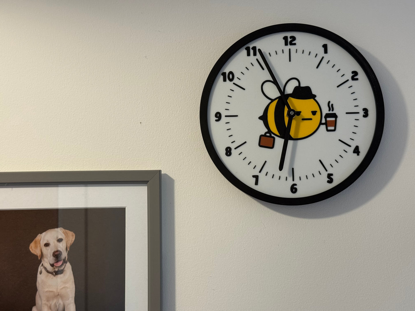 Not Made For This Wall clock with a cartoon design of a bee and a dog, next to a framed picture of a dog on a white wall.