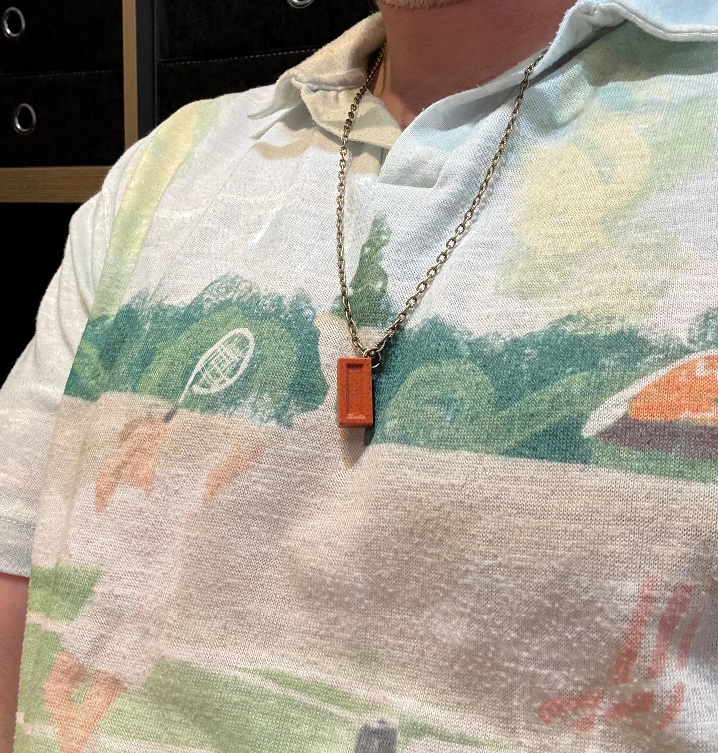 Person wearing a necklace with a rectangular pendant, standing in front of a blurred background.