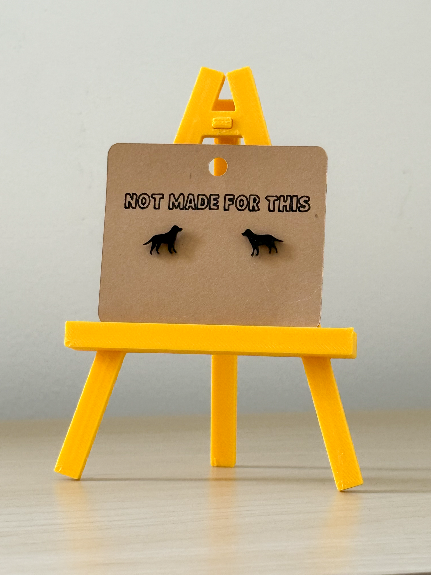 Pair of earrings on a card with 'Not Made For This' text on a yellow stand against a gray background.