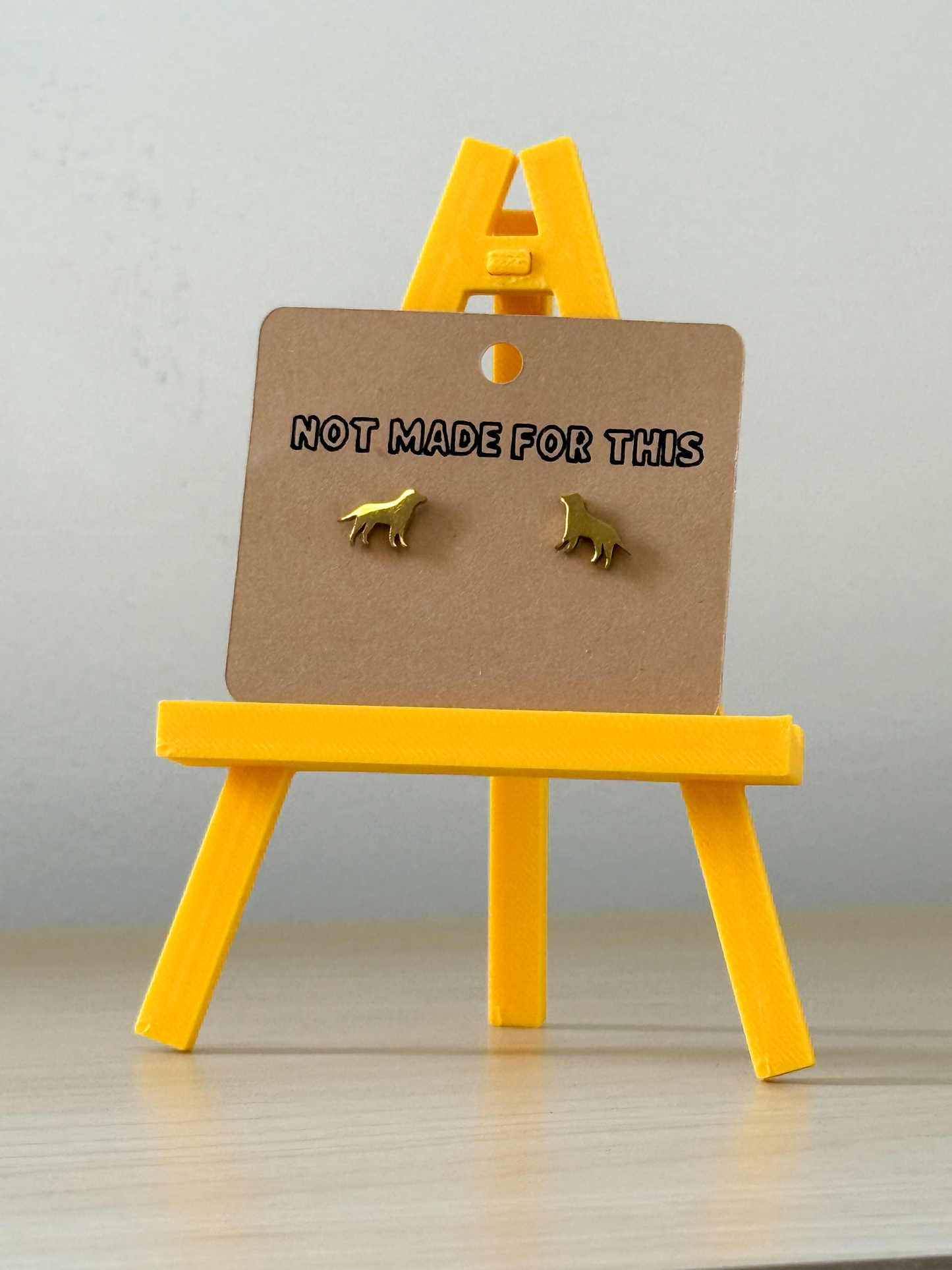 Gold dog-shaped earrings on a brown card with 'Not Made For This' text, displayed on a yellow stand against a light gray background.