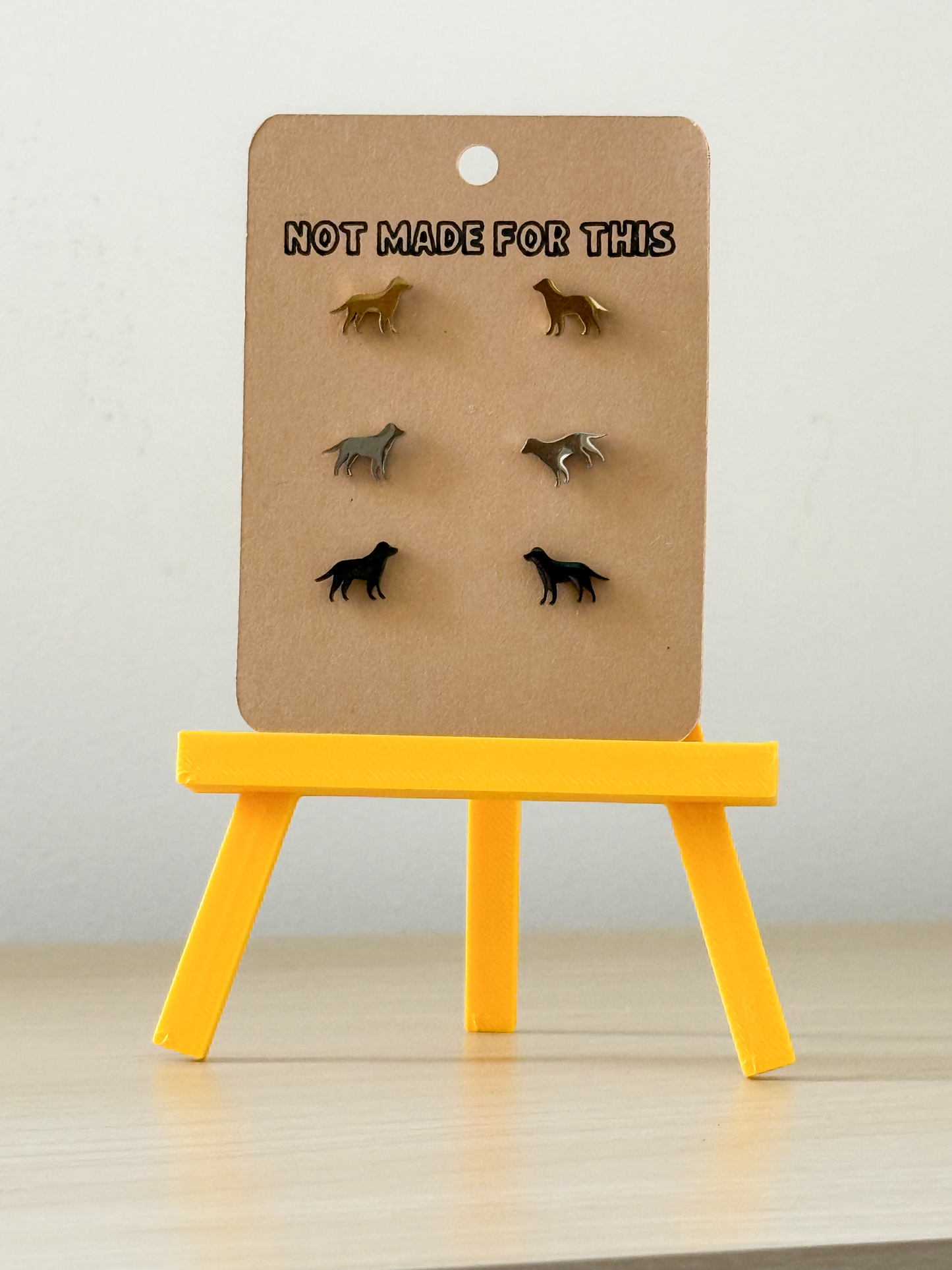 Set of dog-shaped earrings on a cardboard stand with 'Not Made For This' text, placed on a yellow stand against a light background.