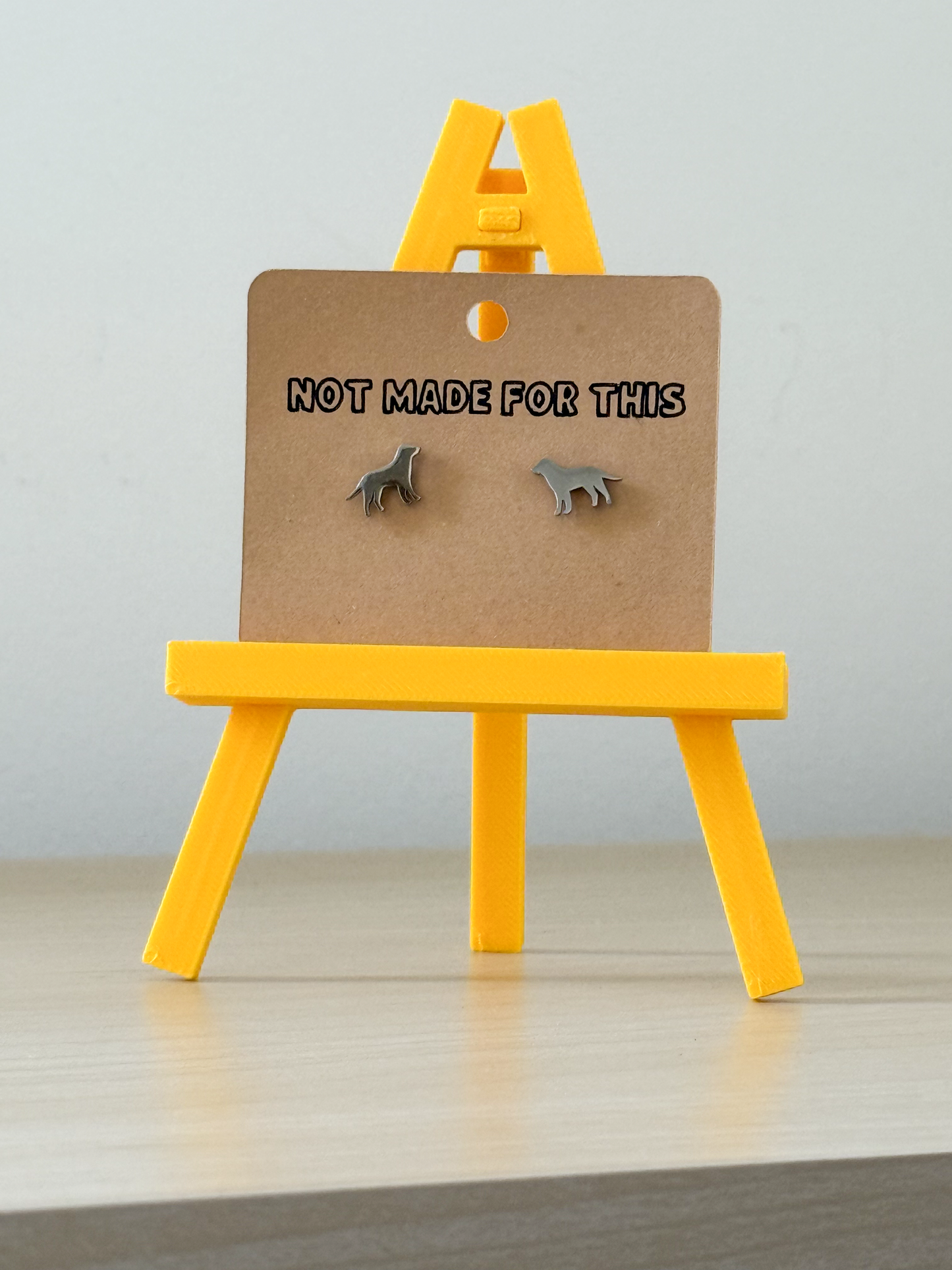 Small dog earrings on a card with 'Not Made For This' text, displayed on a yellow stand against a light background.
