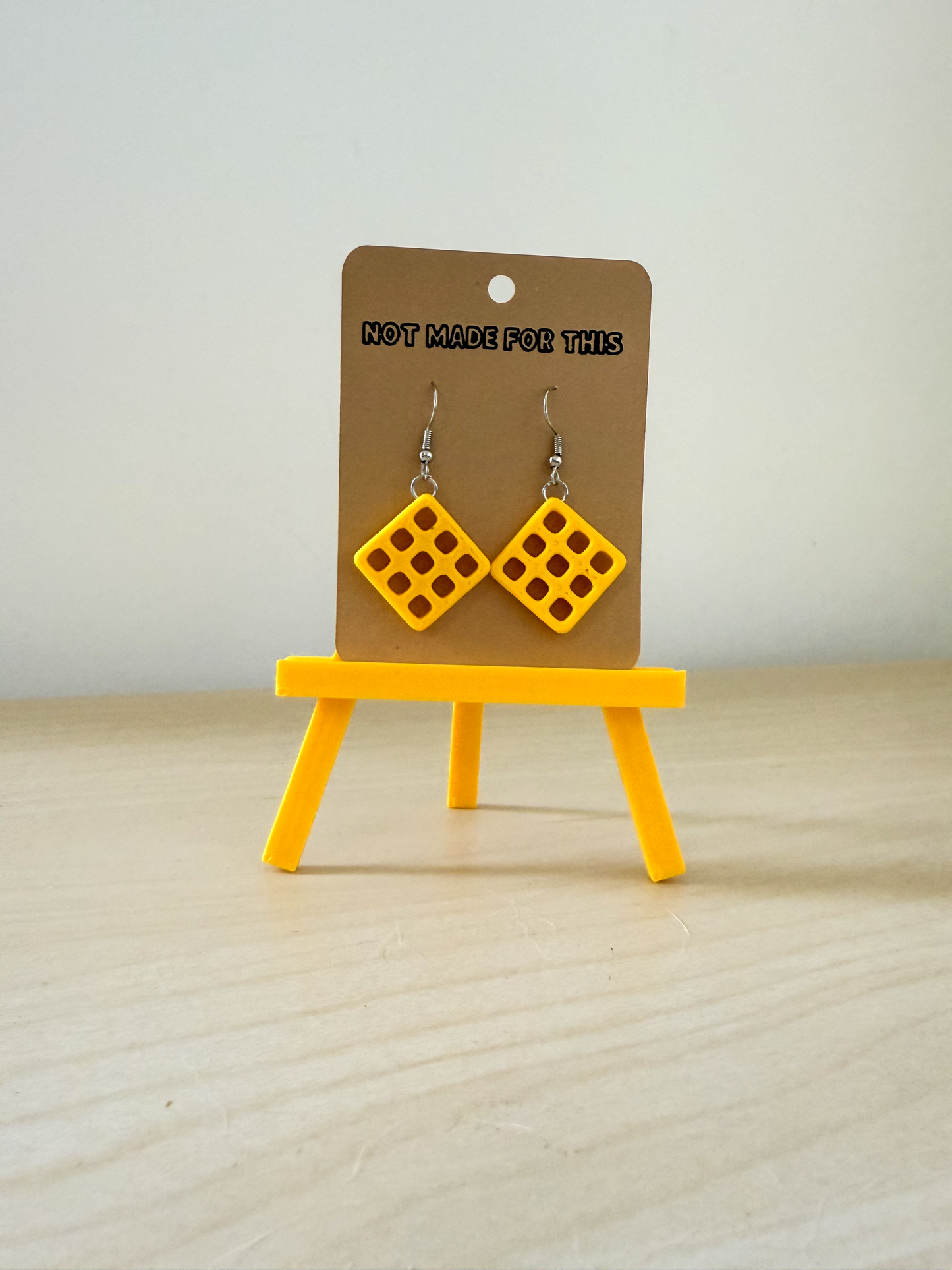 Yellow Potato Waffle earrings on a brown card with 'Not Made For This' text, displayed on a small yellow easel.