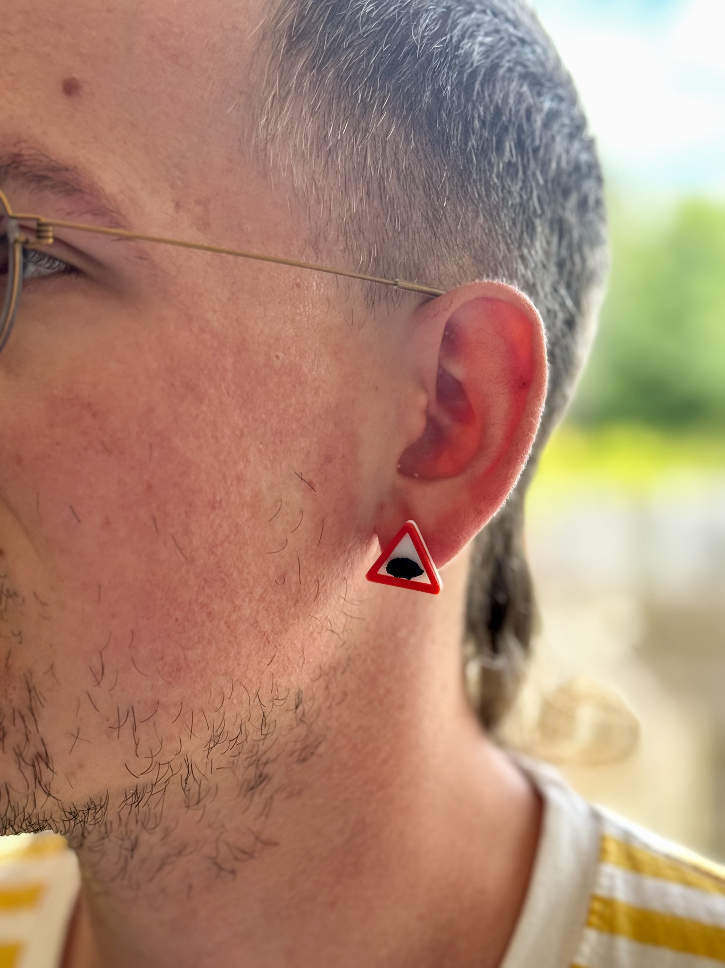 Close-up of a person's ear with a Not Made For This triangular earring, outdoors.