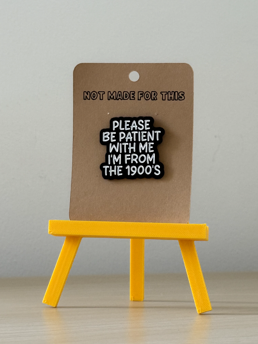 Pin with text 'Please be patient with me I'm from the 1900's' on a brown card with 'Not made for this' text, displayed on a yellow stand against a gray background.