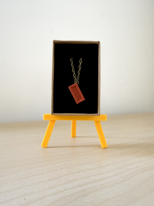 Necklace with a red brick on a black card, displayed on a small yellow easel.