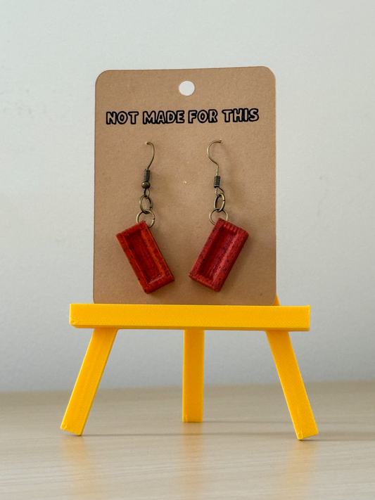 Red earrings on a brown card with 'Not Made For This' text, displayed on a yellow stand.