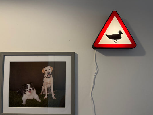 Framed picture of two dogs on a wall next to a triangular warning sign with a duck symbol made by Not Made For This