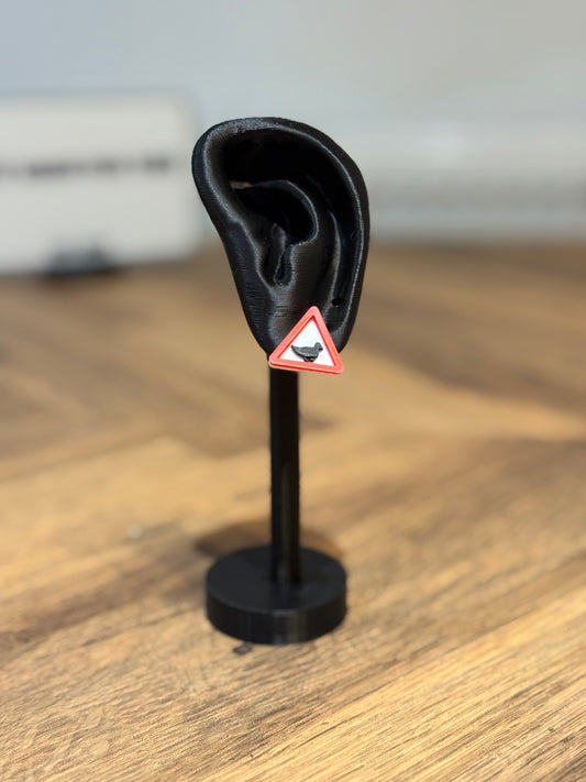 Black ear-shaped object on a stand with a warning triangle duck earring made by Not Made For This on a wooden surface.