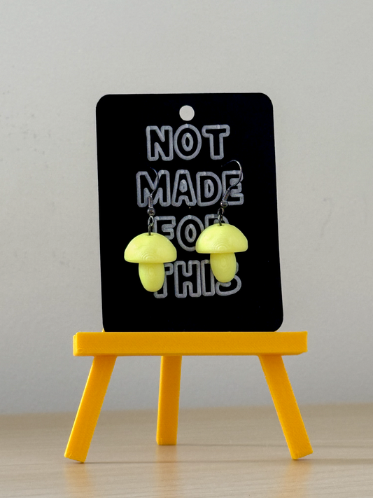 Yellow mushroom-shaped earrings on a black card with 'Not Made for This' text, displayed on a small yellow stand.