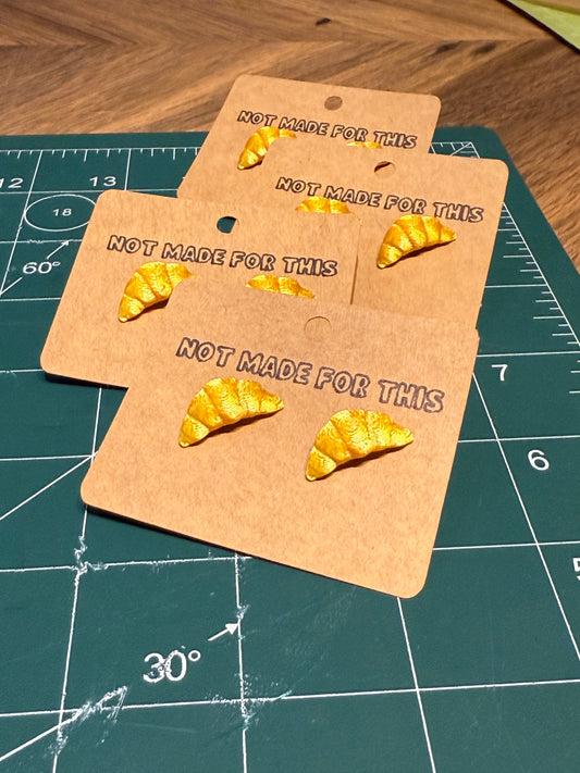 Gold croissant Not Made For This earrings on a 'Not Made For This' card on a green cutting mat.