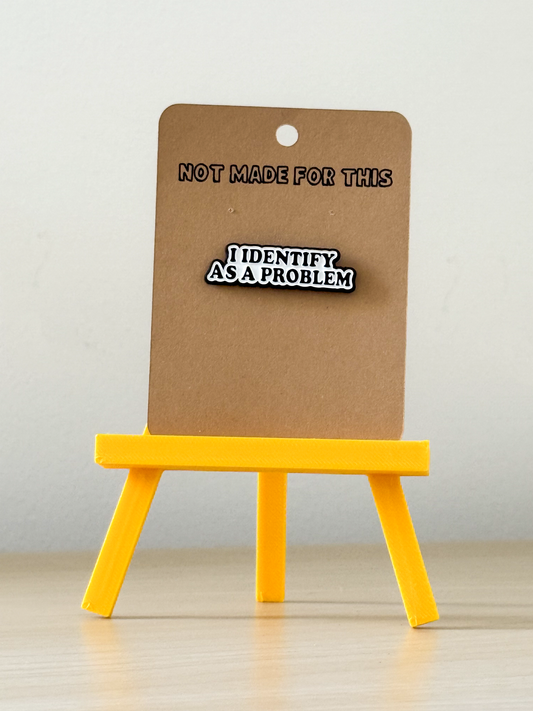 Enamel pin labeled 'I Identify as a Problem' on a cardboard stand with text 'Not Made for This'.