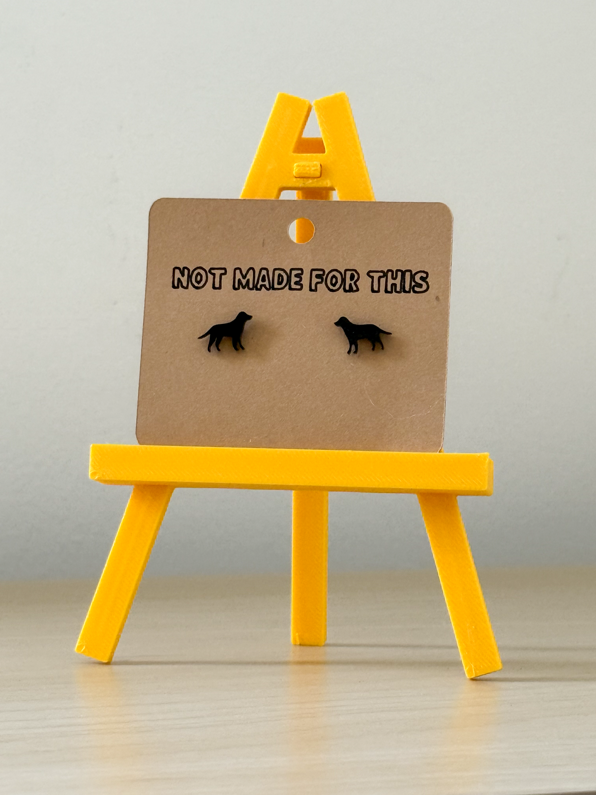 Pair of earrings on a card with 'Not Made For This' text on a yellow stand against a gray background.