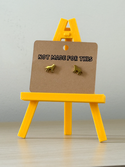 Gold dog-shaped earrings on a brown card with 'Not Made For This' text, displayed on a yellow stand against a light gray background.