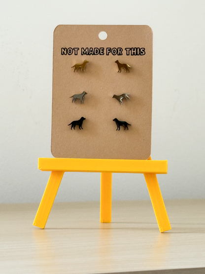 Set of dog-shaped earrings on a cardboard stand with 'Not Made For This' text, placed on a yellow stand against a light background.
