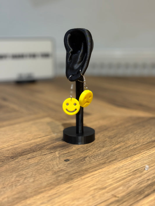 Not Made For This  Yellow smiley face earrings on a black stand resembling an ear, on a wooden surface.