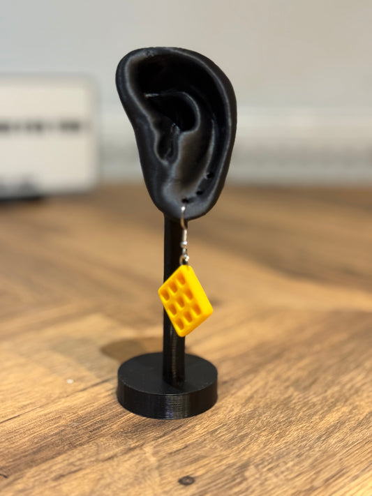 Yellow Not Made For This waffle-shaped earring on a black ear stand with a blurred background