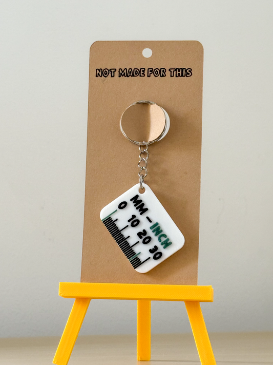 Keychain with measurement scale on a cardboard backing with 'Not Made For This' text, placed on a yellow stand.