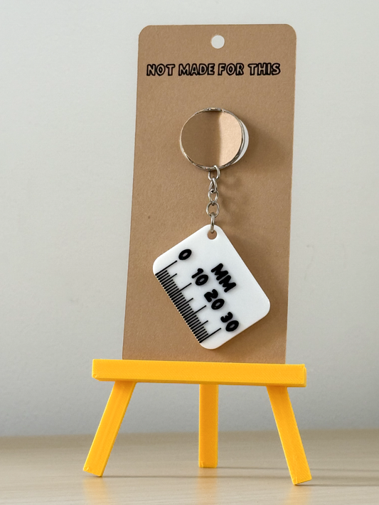 Keychain with a ruler design on a brown card, placed on a yellow stand against a gray background.
