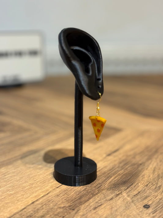 Not Made For This Pizza slice earring on a black stand shaped like an ear on a wooden surface.