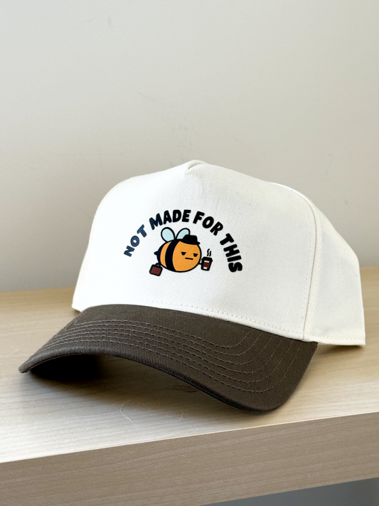 White cap with brown bill featuring a bee graphic and Not Made For This text on a light wooden surface.